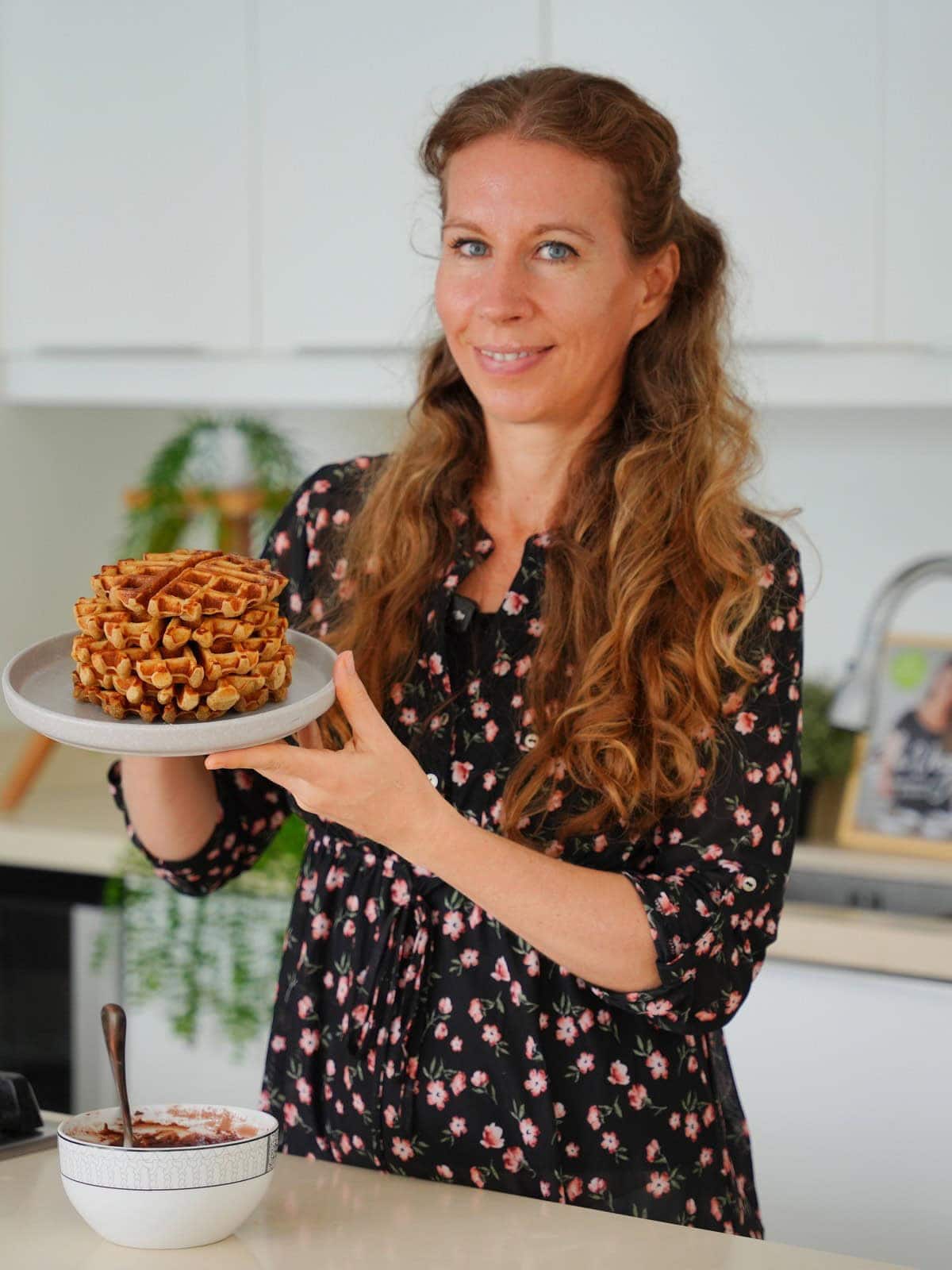 Michaela Vais in a bright kitchen smiling and holding a stack of waffles