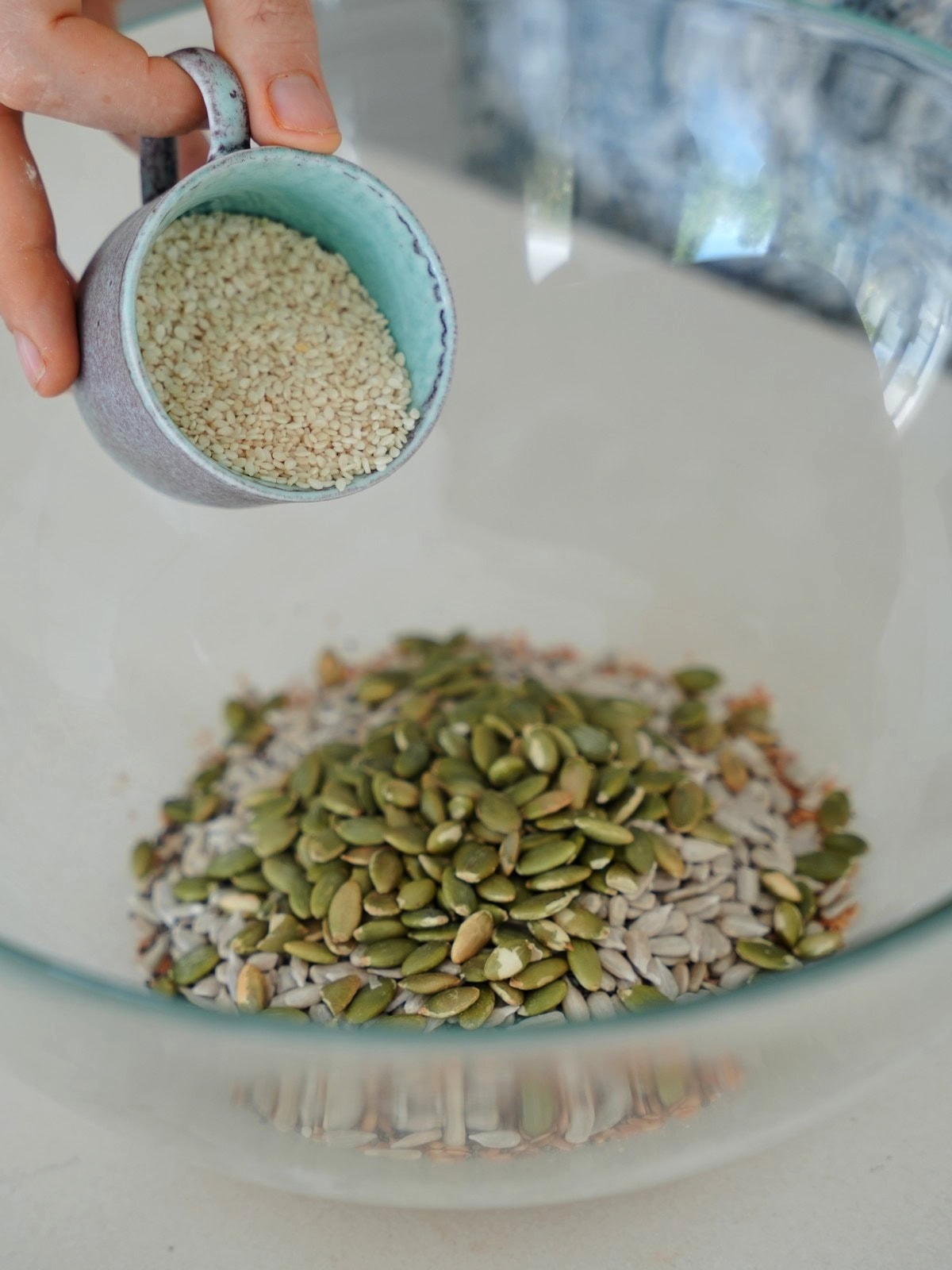 A hand holding a small cup of sesame seeds above a glass bowl filled with pumpkin and sunflower seeds