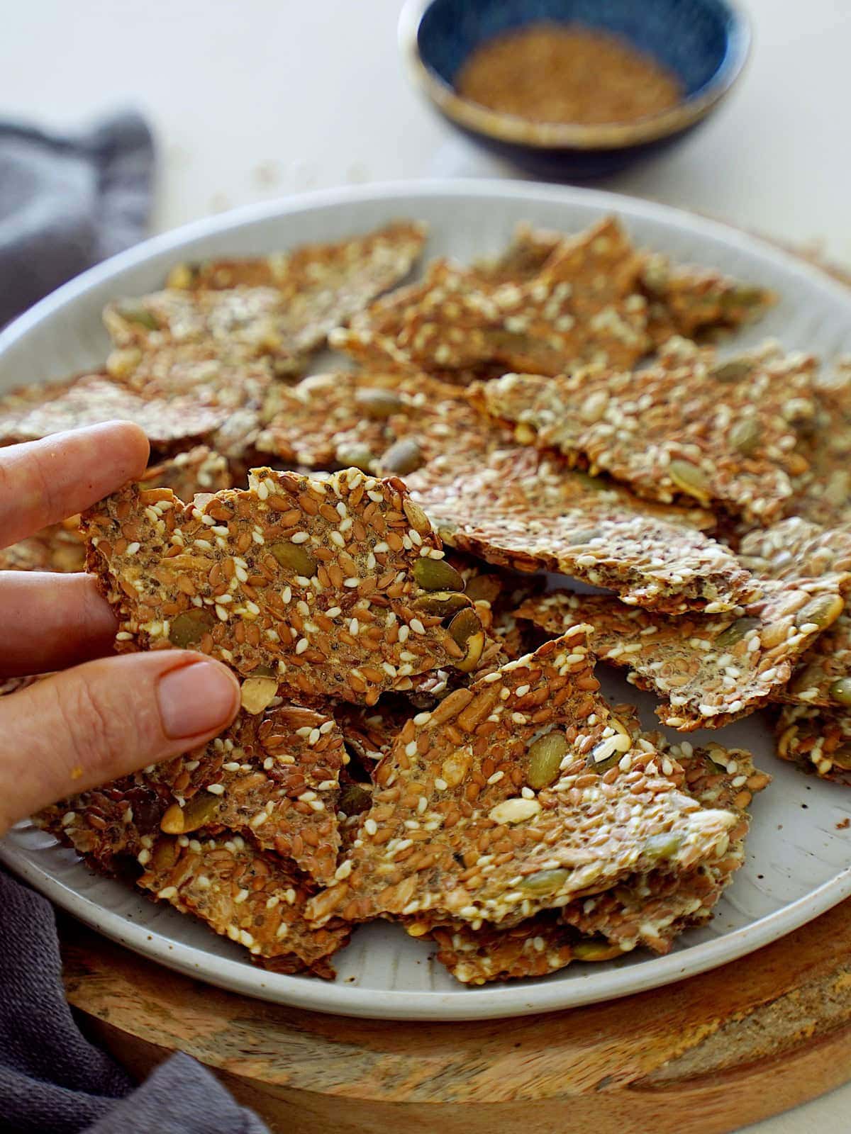 A hand reaching for a crispy seed cracker on a white plate surrounded by more crackers