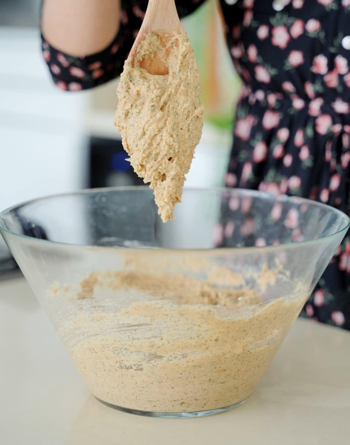 A person holding a wooden spoon with thick, creamy batter above a glass mixing bowl filled with similar batter