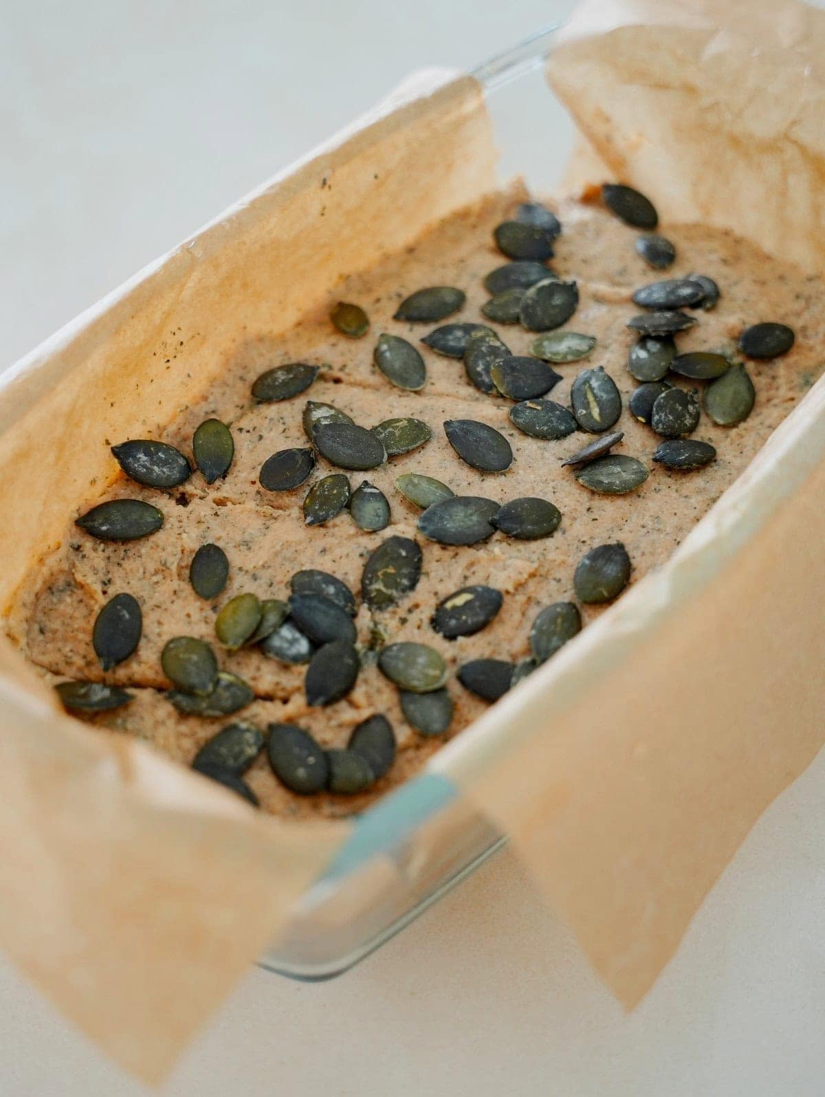 Light brown batter topped with dark green pumpkin seeds, placed in a glass container lined with parchment paper.