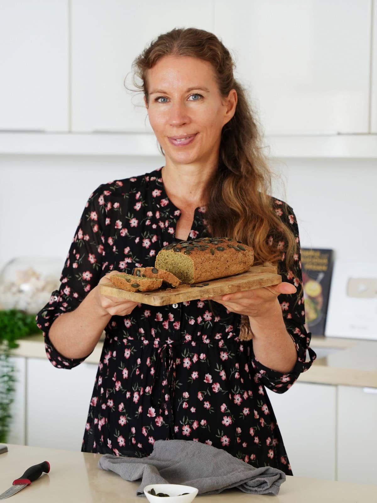 Michaela Vais in a floral blouse holds a loaf of lentil bread on a wooden board in a modern kitchen.