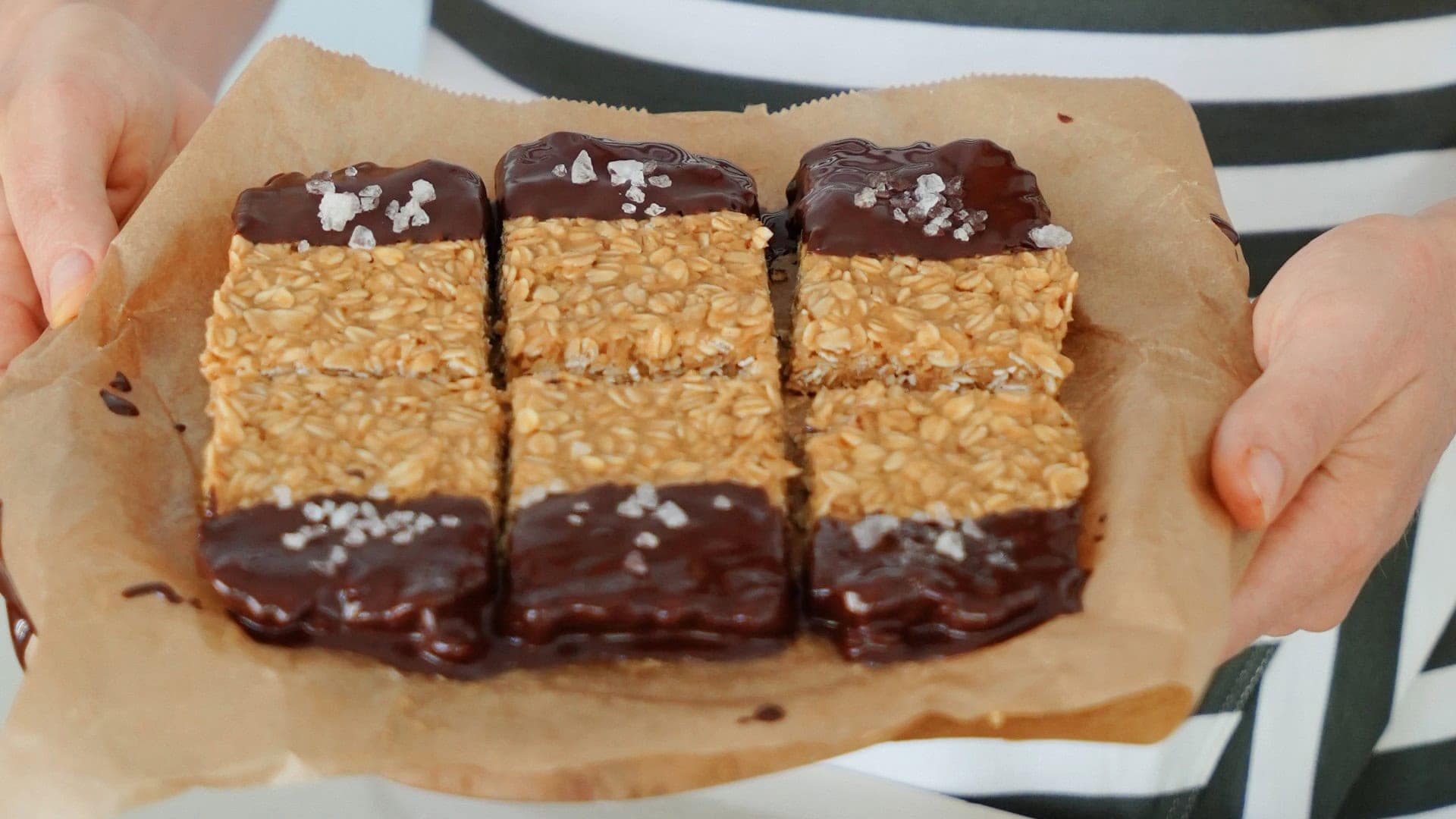 hands hold a parchment paper lined plate with 6 peanut butter oatmeal bars dipped in chocolate.