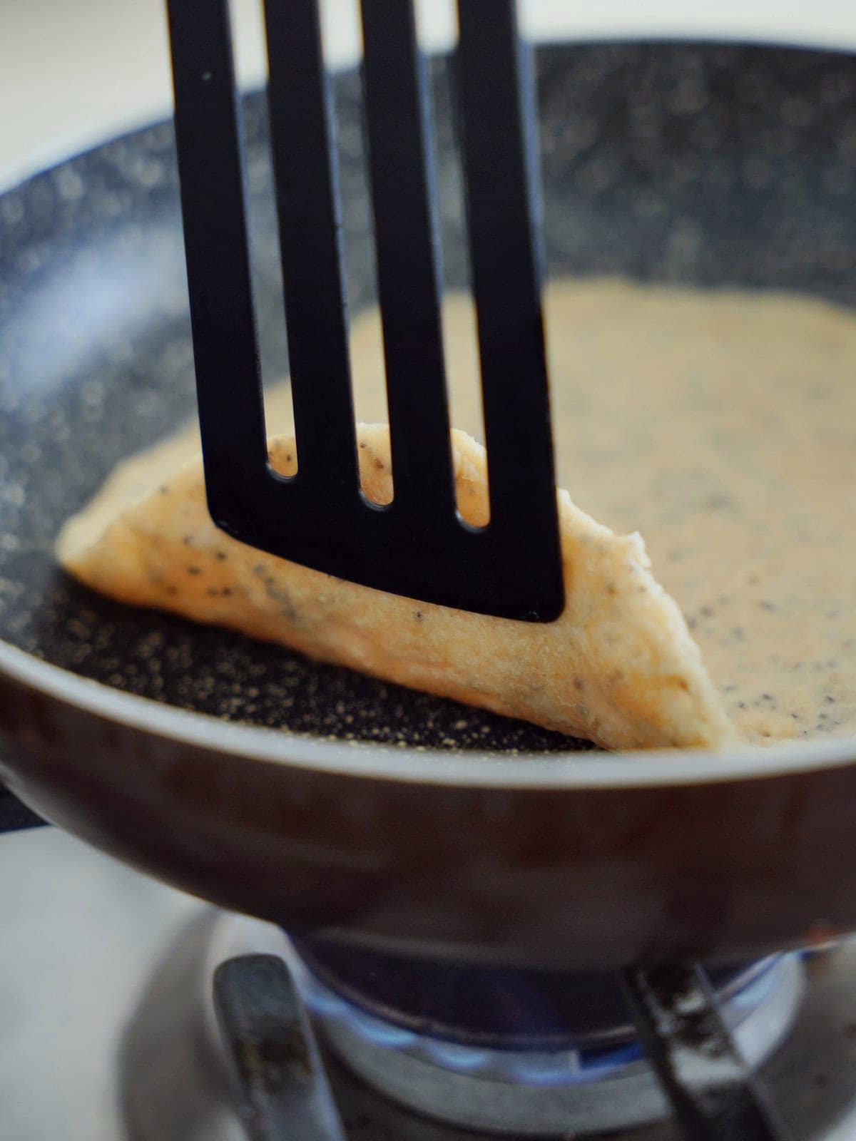 A black spatula lifts the edge of a thin, speckled pancake cooking in a frying pan.