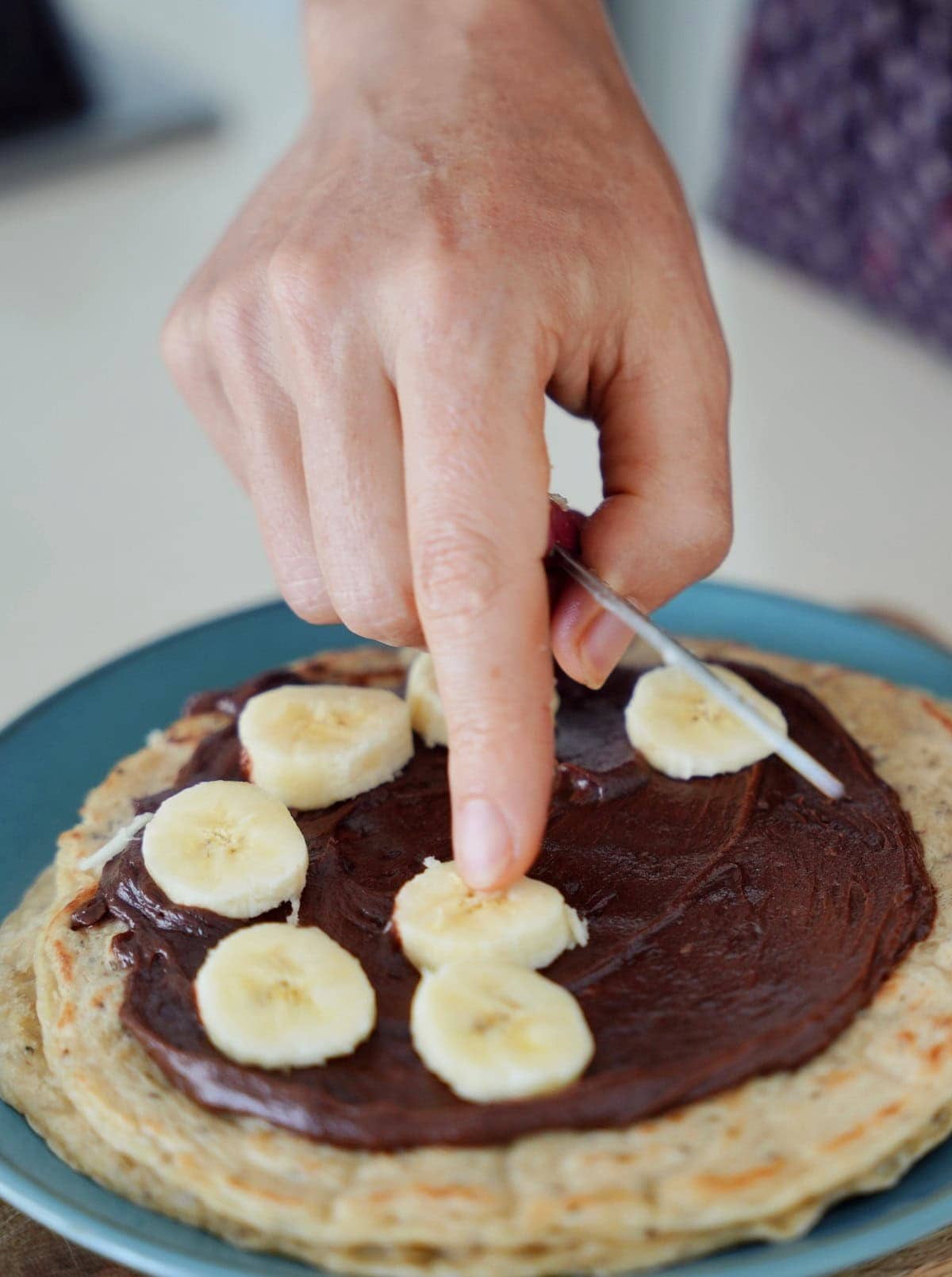 A hand places a slice of banana onto a chia oat wrap with chocolate cream