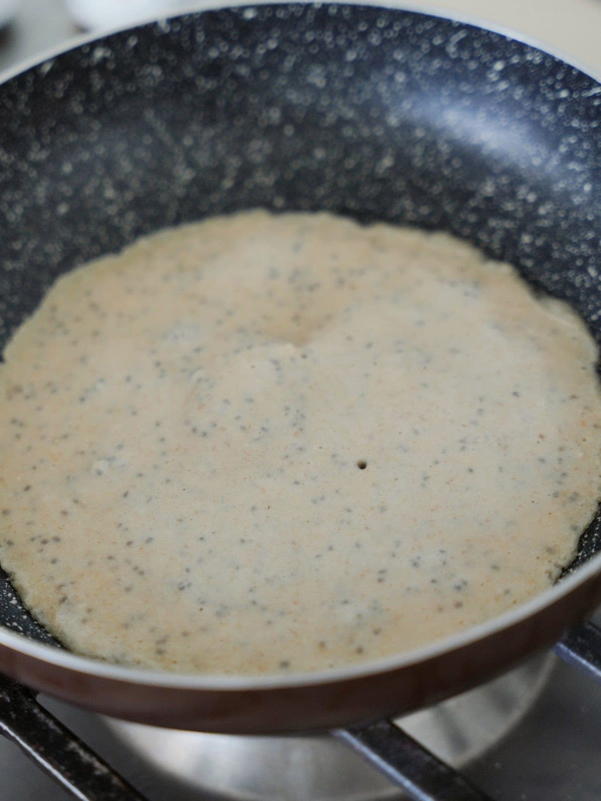 A thin, round pancake batter with visible seeds cooking in a frying pan.